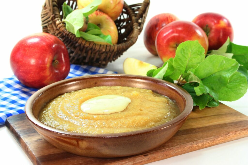 A bowl of applesauce next to apples and herbs.