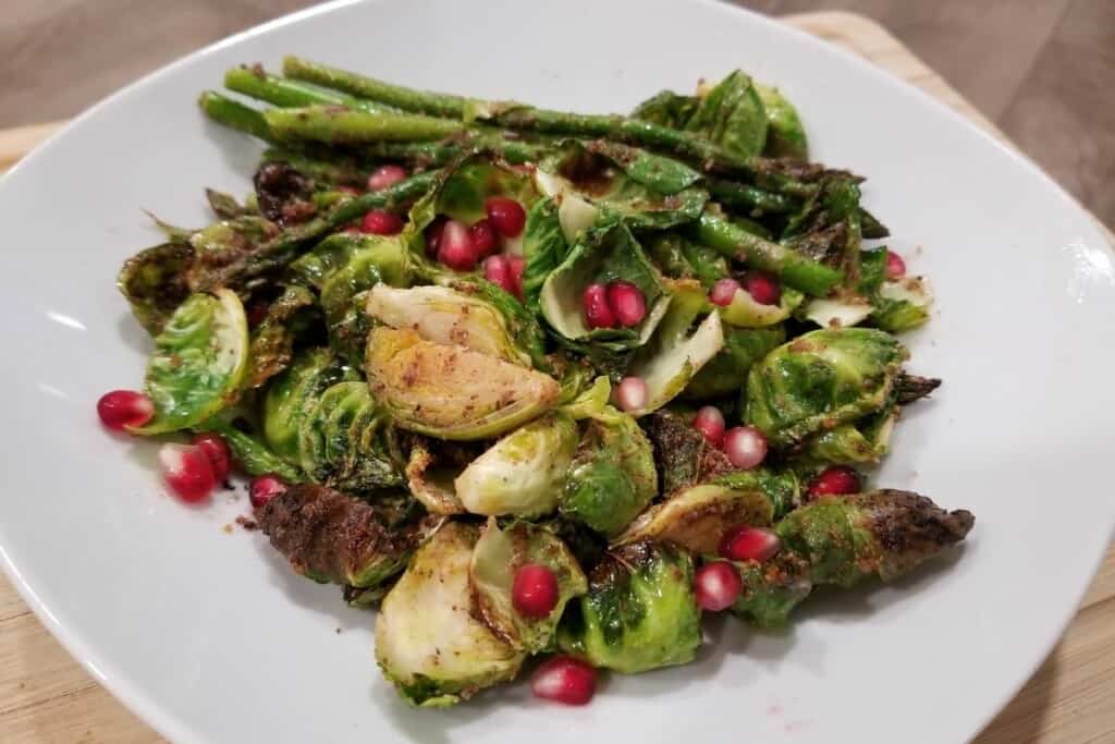 roasted, seasoned asparagus and brussels sprouts on a white plate topped with pomegranate seeds.