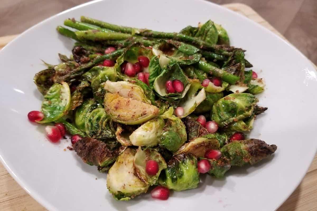 roasted, seasoned asparagus and brussels sprouts on a white plate topped with pomegranate seeds.