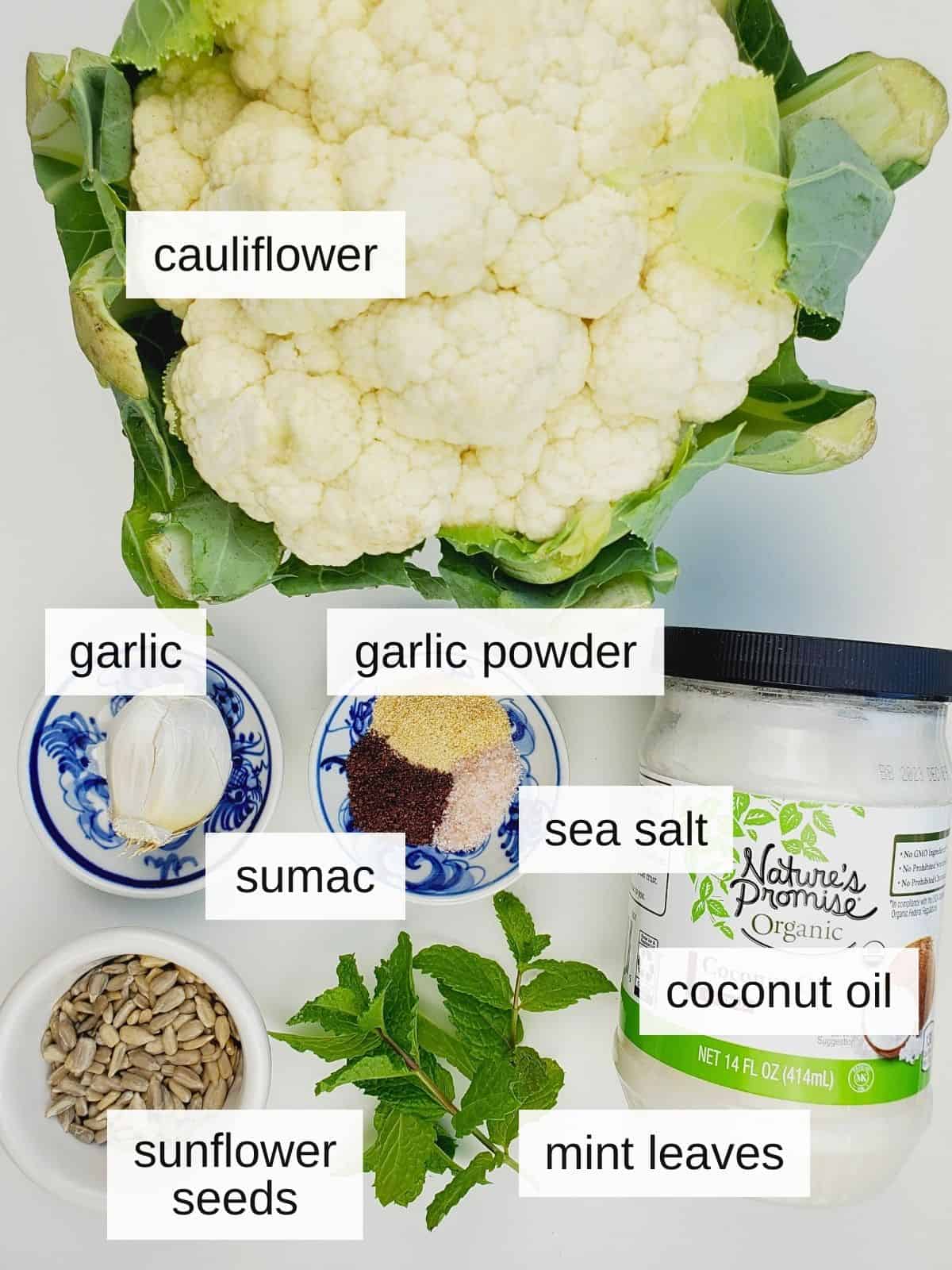 ingredients for air fryer whole cauliflower, including whole cauliflower, garlic cloves, garlic powder, sumac, sea salt, coconut oil, sunflower seeds, and mint leaves.