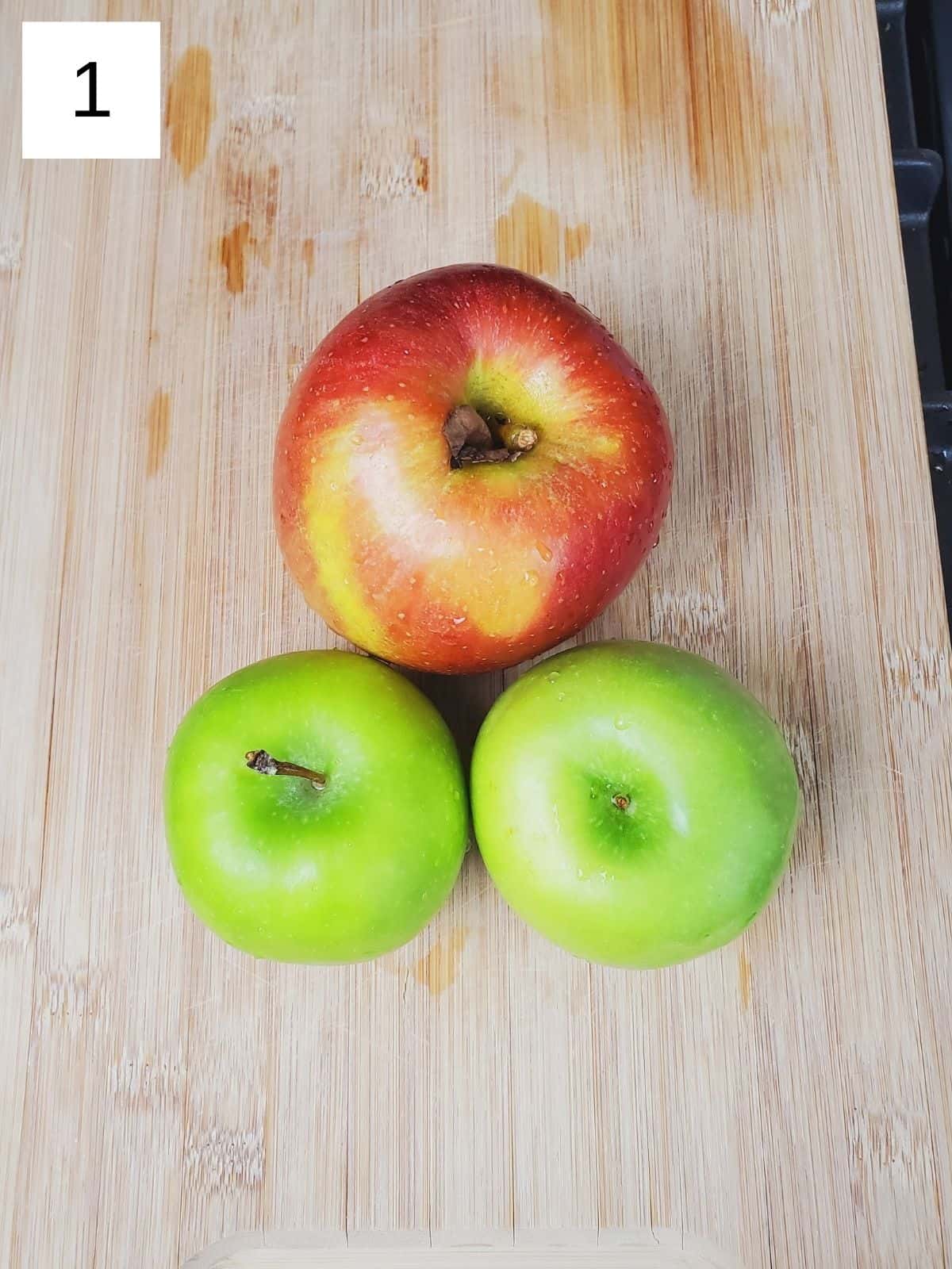 three medium-sized apples on a wooden cutting board.