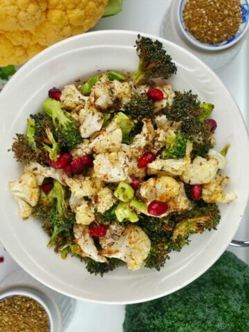 cooked seasoned broccoli and cauliflower, topped with pomegranate seeds, on a white plate.