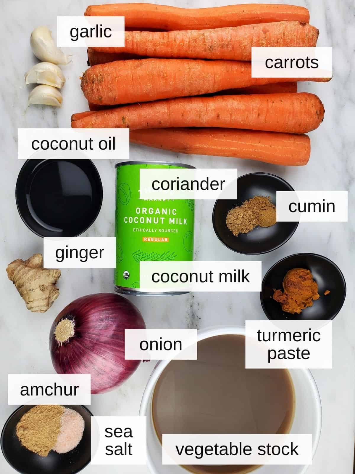 ingredients for carrot ginger coconut soup, including red onion, coconut milk, ginger, turmeric, coriander, cumin, sea salt, amchur powder, garlic, coconut oil, and carrots.