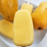 mango popsicles and two fresh mangoes on a white plate.