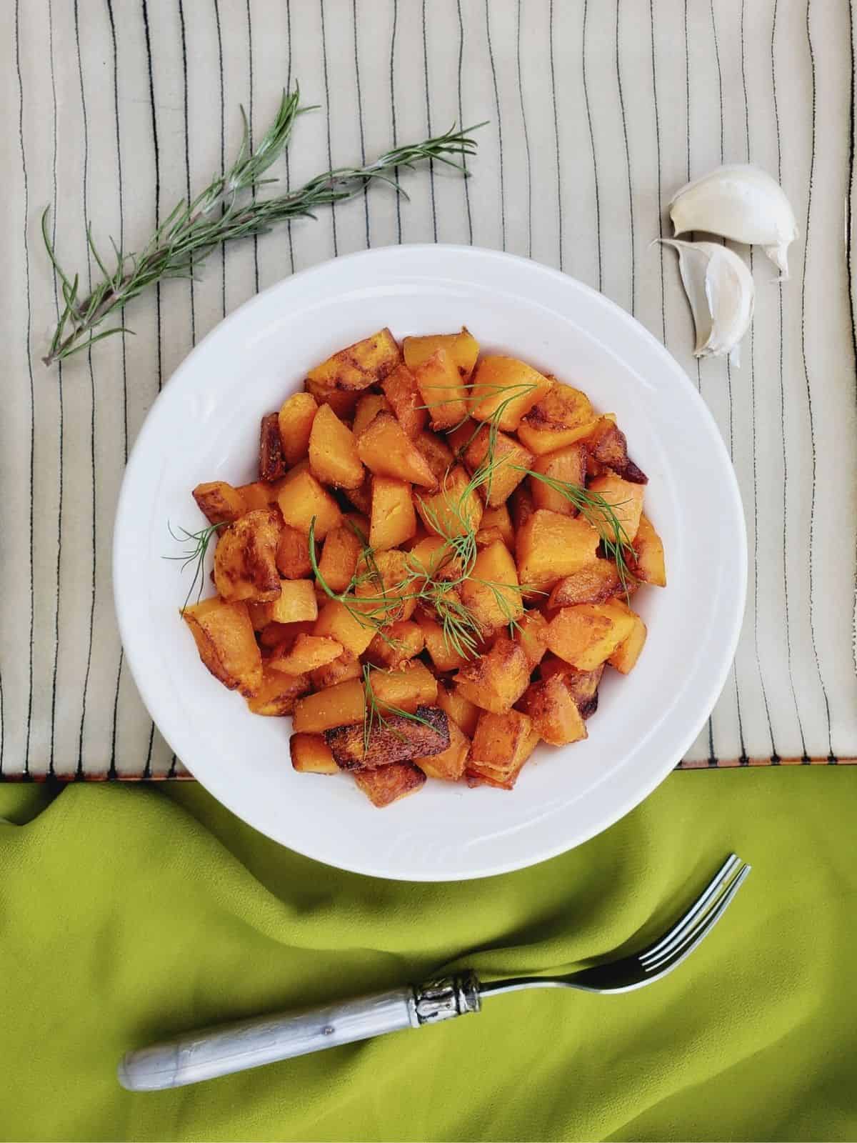 seasoned and sautéed butternut squash pieces, topped with fresh rosemary, on a white plate.