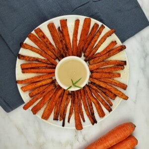 air fried carrots fries arranged in a concentric circle on a white plate around a small container of sauce.