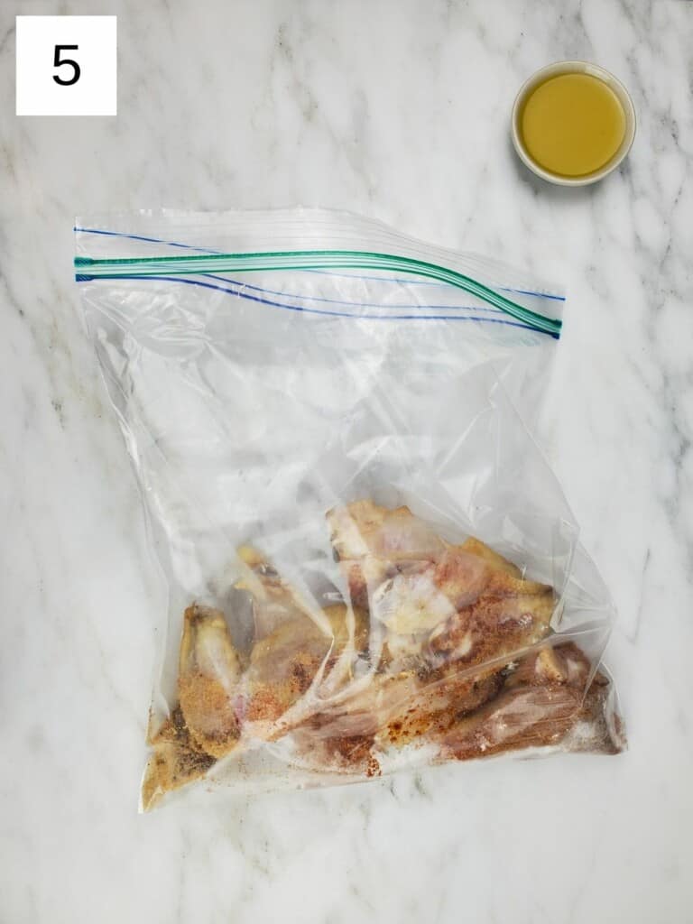 ziploc bag of chicken wings with spices and a small bowl of oil, on top of a marble slab.