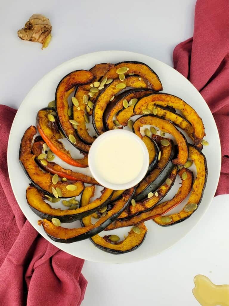 slices of roasted acorn squash arranged in a concentric circle on a large white plate around a small container of white sauce.