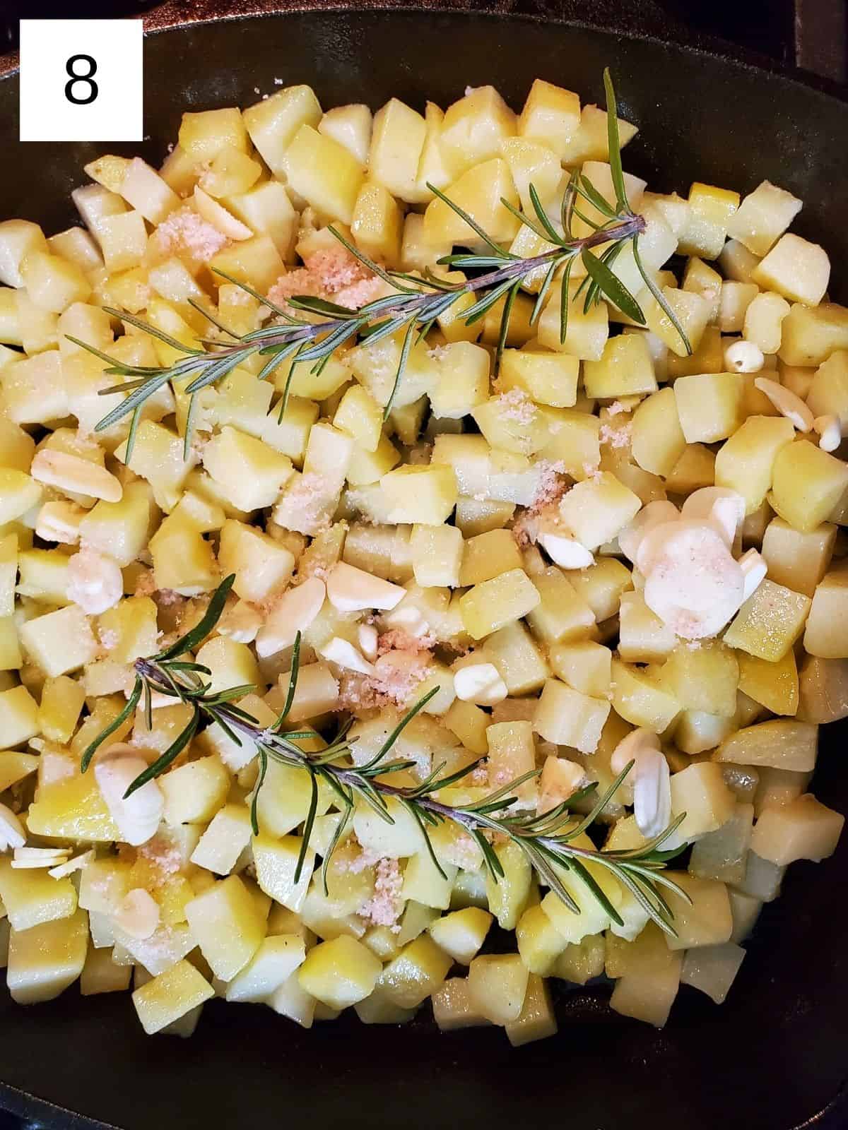 potato cubes, topped with seasonings and rosemary sprigs, in an oil-coated cast iron pan.