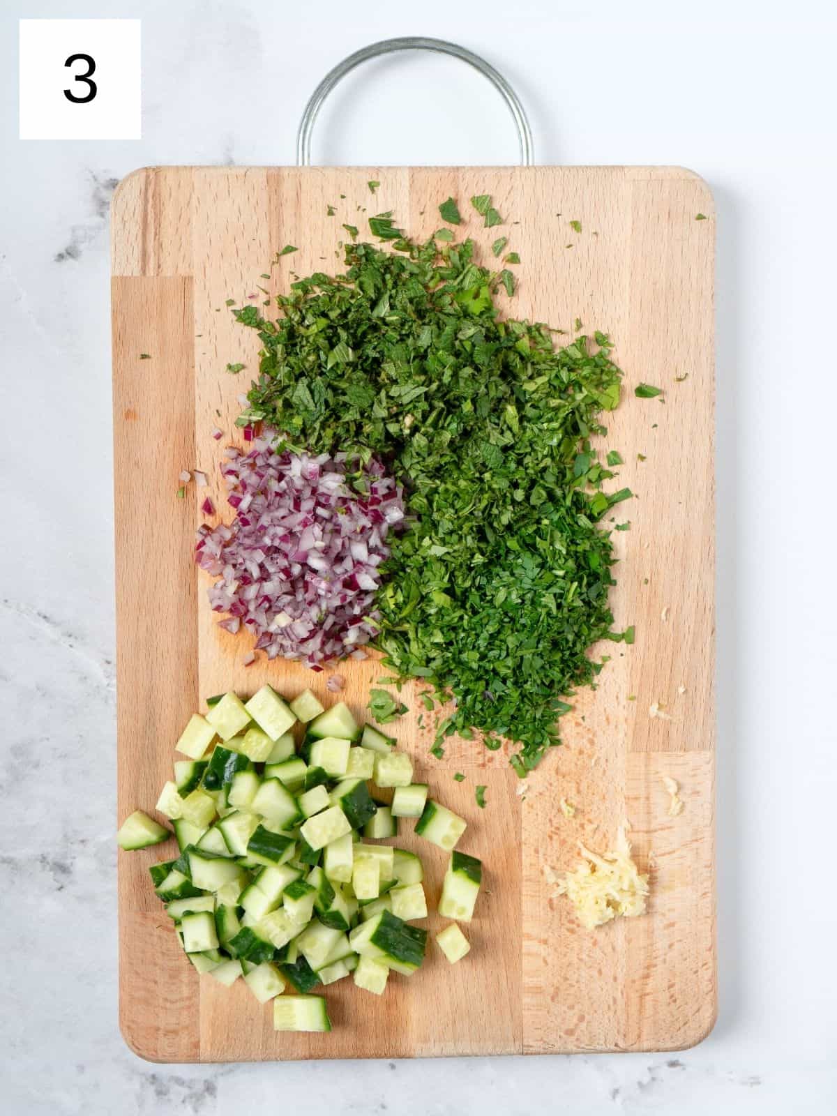 finely chopped various herbs and spices, and cucumber on a wooden chopping board.