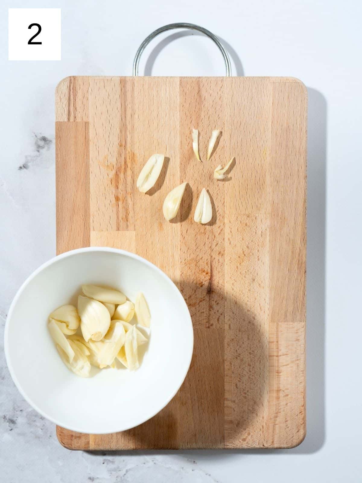 a bowl of peeled garlic cloves and some sliced garlic with its green sprouts removed, on a wooden chopping board.