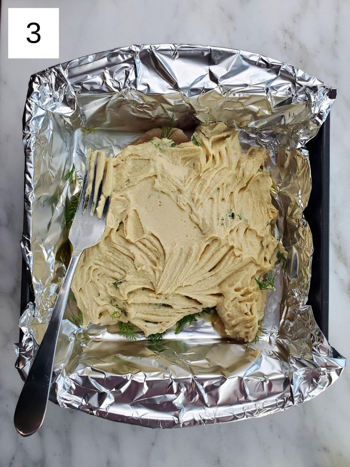chicken tenders, topped with seasonings and hummus, arranged on a foil-lined baking tray.