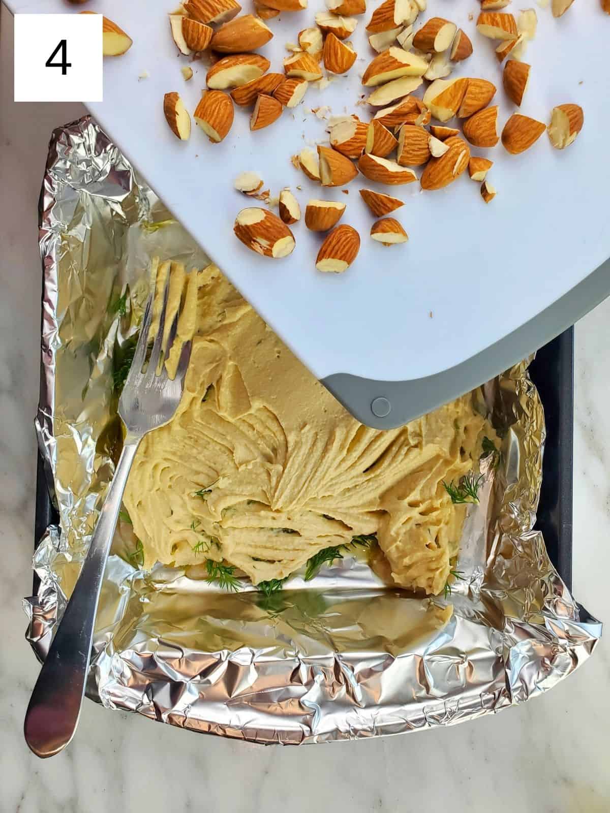 chopped almonds being sprinkled over hummus-topped chicken tenders on a foil-lined baking tray.