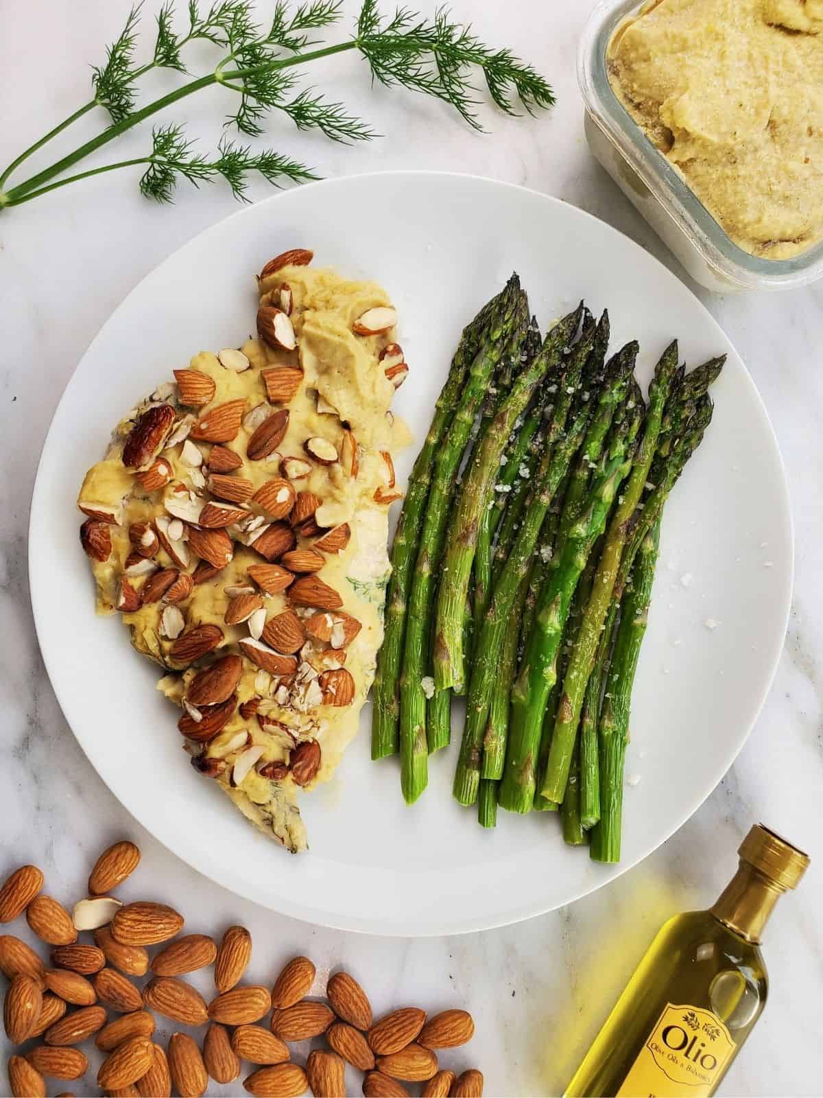 a plate of baked chicken covered in hummus, served with cooked asparagus.