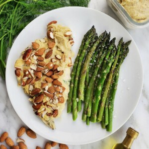 a plate of baked chicken covered in hummus, served with cooked asparagus.