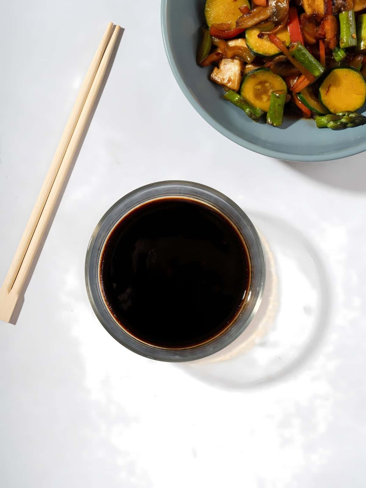 A small sauce bowl of vegan stir-fry sauce next to a bowl of fried vegetables and a pair of chopsticks.