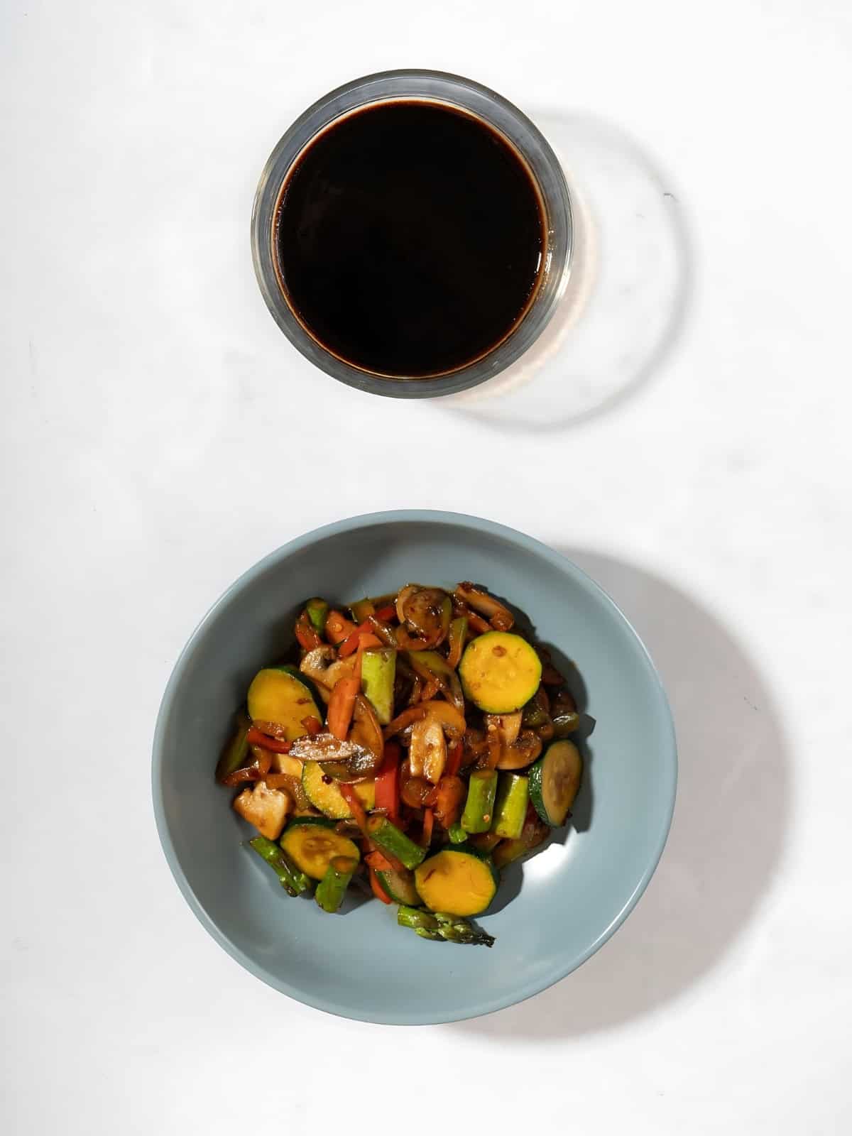 A small sauce bowl of vegan stir-fry sauce next to a bowl of fried vegetables.