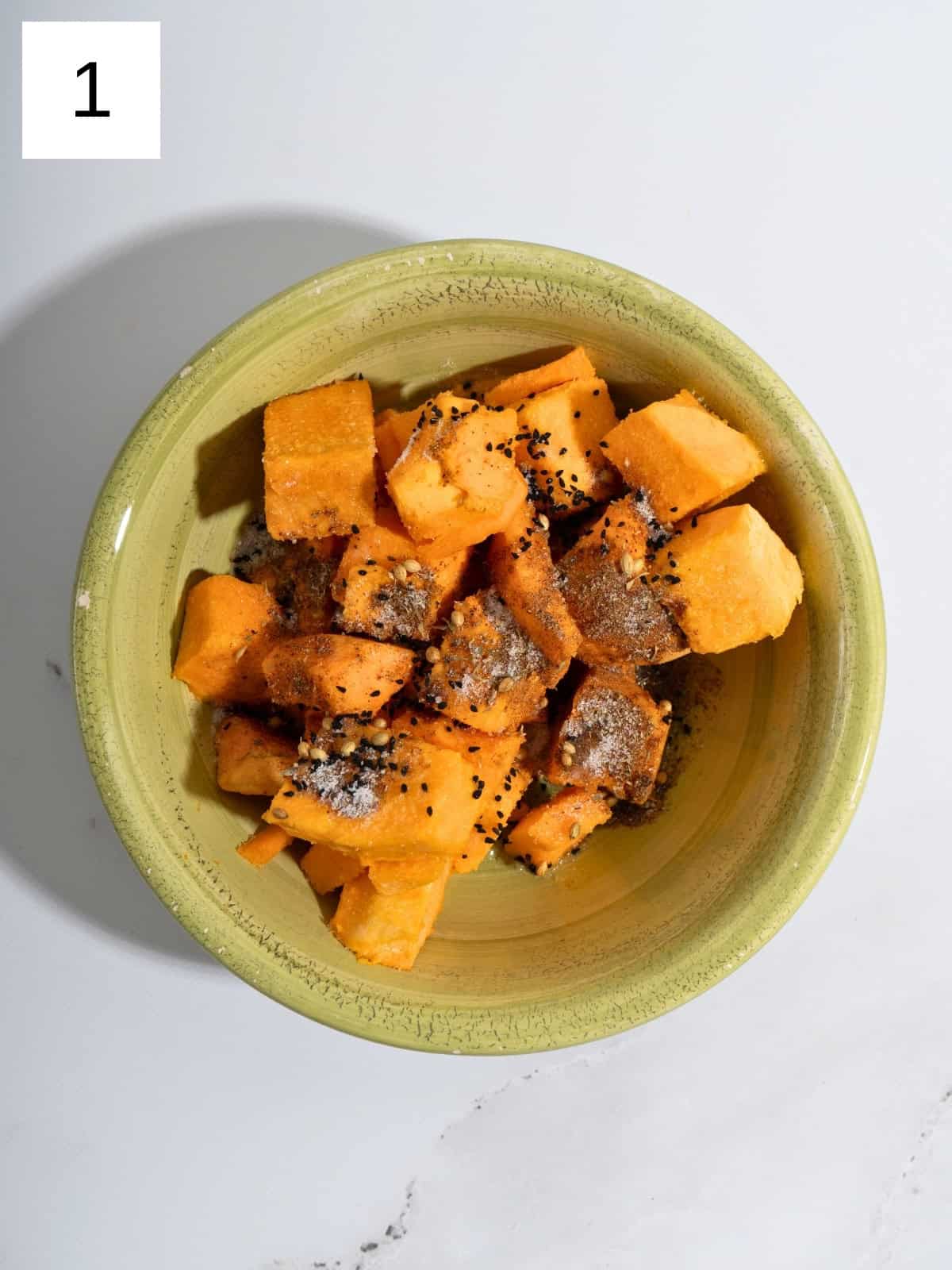 Chopped pieces of butternut squash garnished with oil, za'atar, nigella seeds, sweetener, amchur, and salt in a bowl.