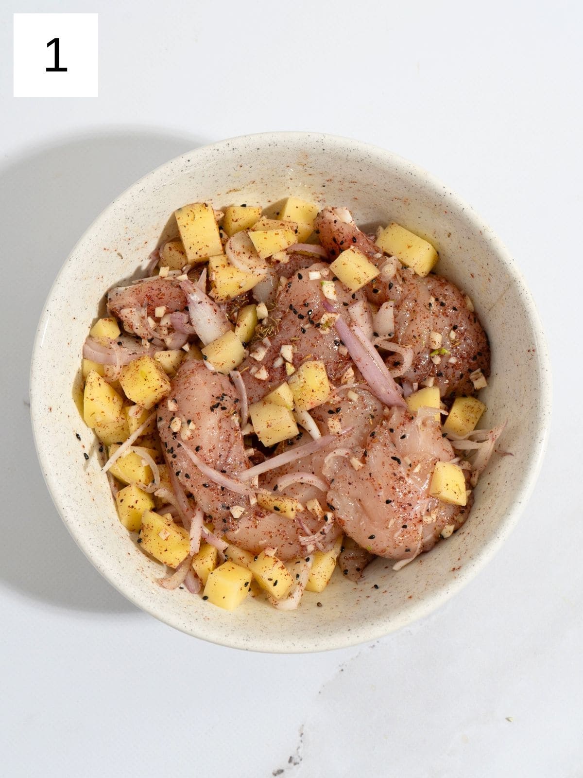 Raw chicken strips, coated with garlic, onion, potato slices, and spices in a bowl.