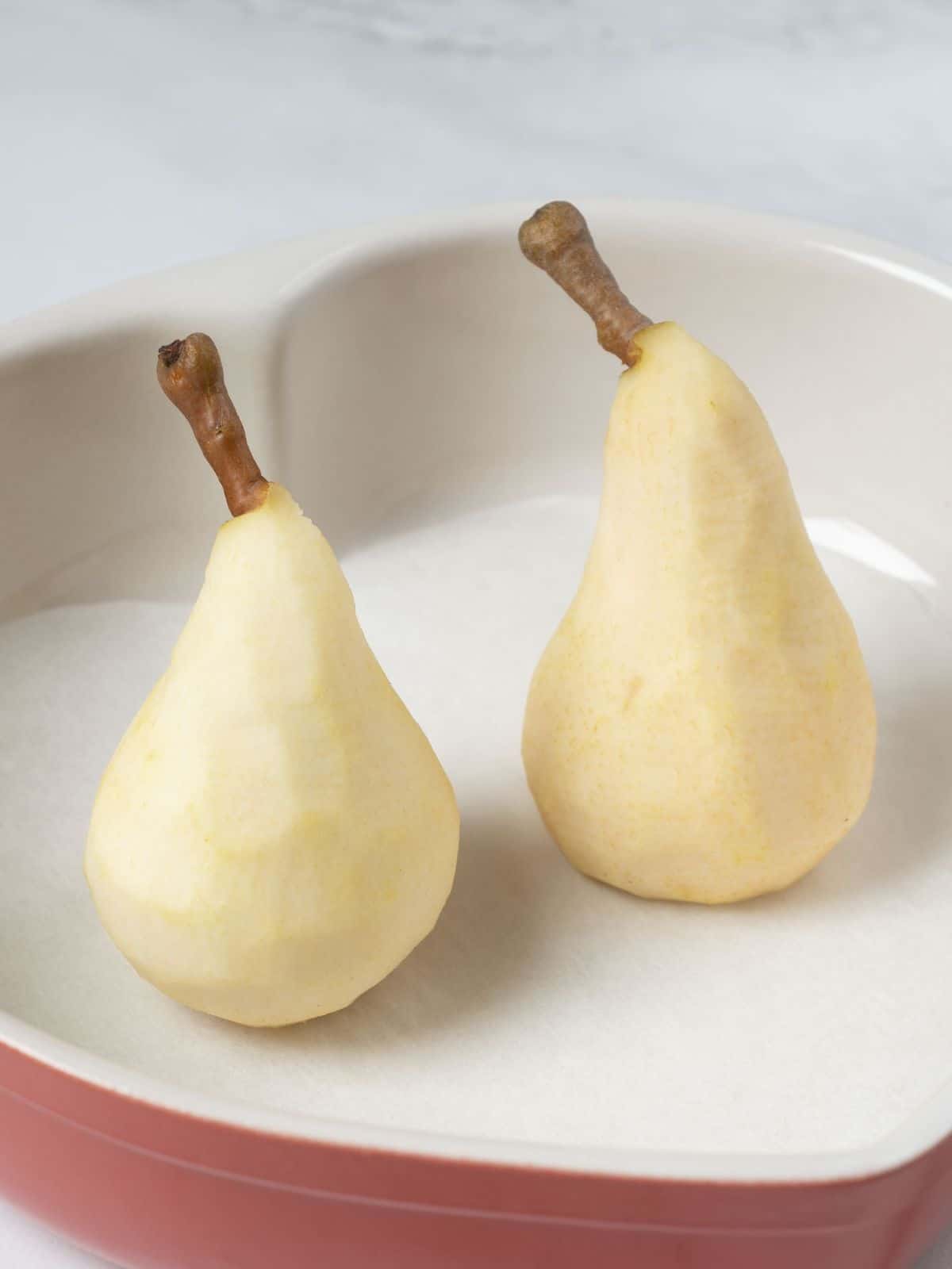 Two peeled pears placed upright in a baking dish, ready for baking, showcasing their smooth texture and inviting shape.

