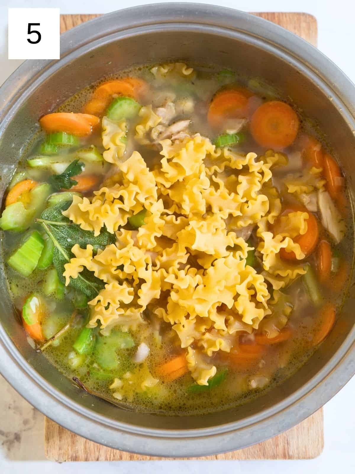 A pot of soup simmering with sliced carrots, celery, and shredded chicken, as noodles are stirred in to cook until tender.