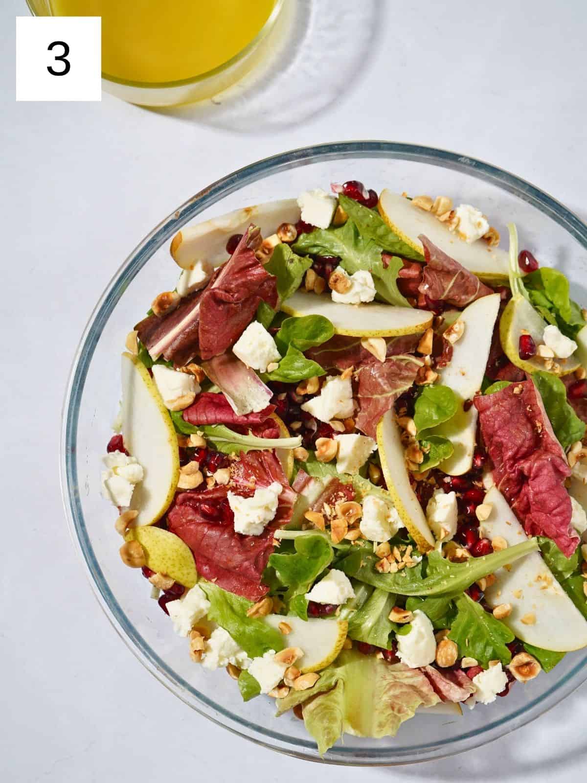 A colorful glass bowl filled with a fresh salad of mixed greens, goat cheese, sweet pear slices, pomegranate seeds, hazelnuts, and a drizzle of apple cider vinegar dressing.