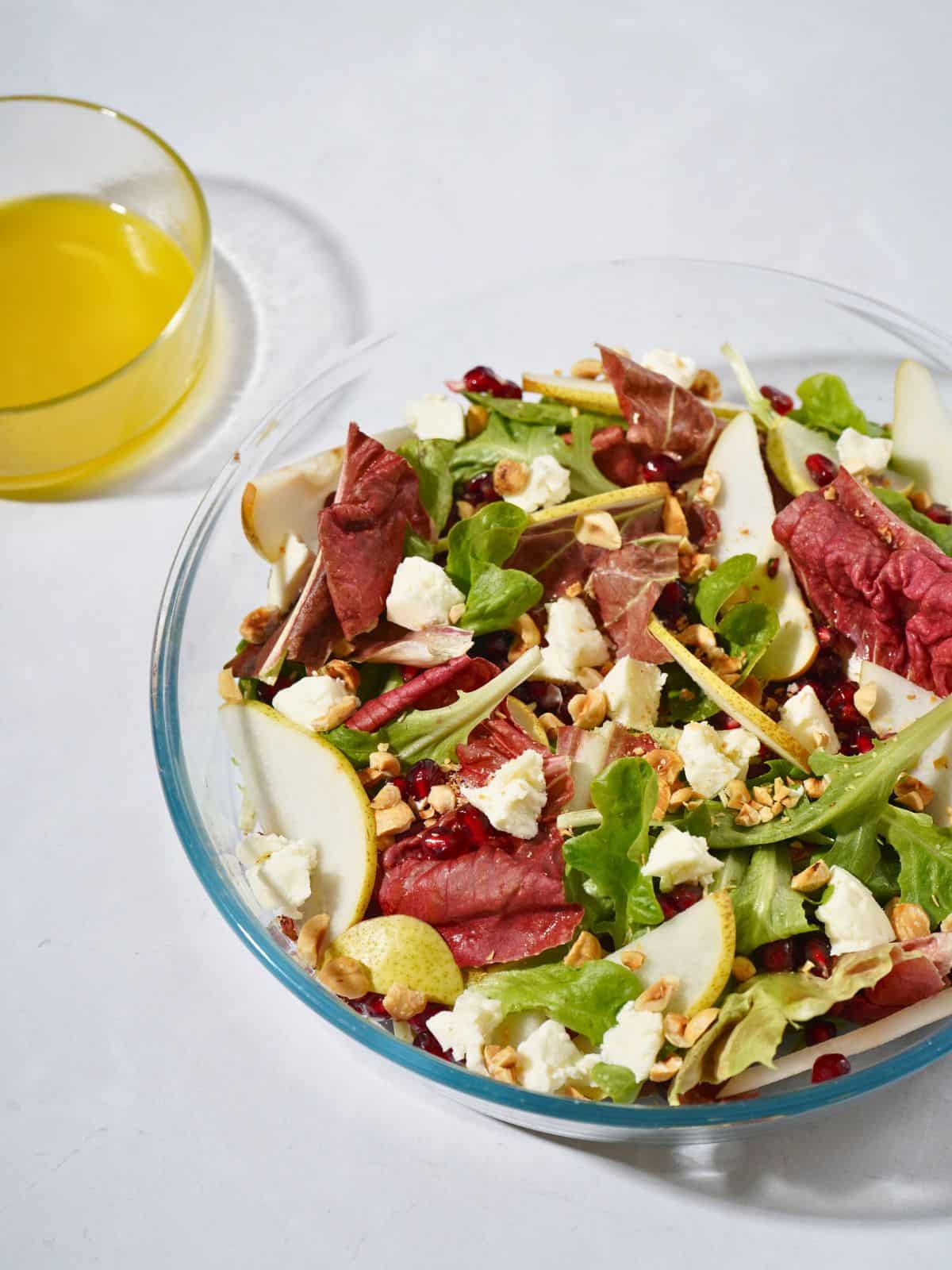 A colorful glass bowl filled with a fresh salad of mixed greens, sweet pear slices, goat cheese, pomegranate seeds, hazelnuts, and a drizzle of apple cider vinegar dressing.