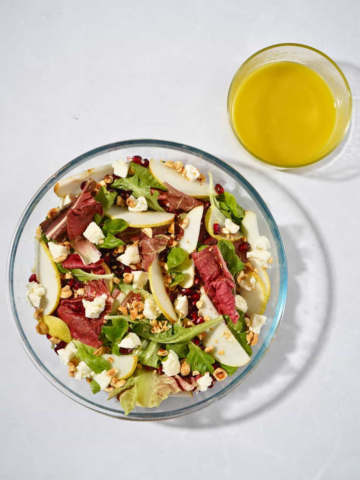A vibrant salad in a glass bowl featuring mixed greens, goat cheese, juicy pomegranate seeds, sliced pears, crunchy hazelnuts, and a tangy apple cider vinegar dressing, beautifully mixed and ready to serve.