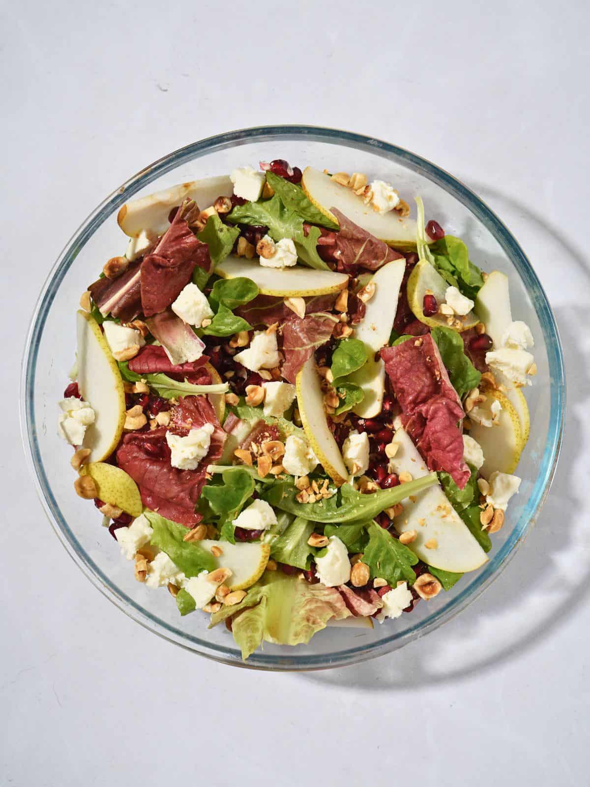 A vibrant salad in a glass bowl featuring mixed greens, goat cheese, juicy pomegranate seeds, sliced pears, and crunchy hazelnuts, perfect for a refreshing and healthy dish.