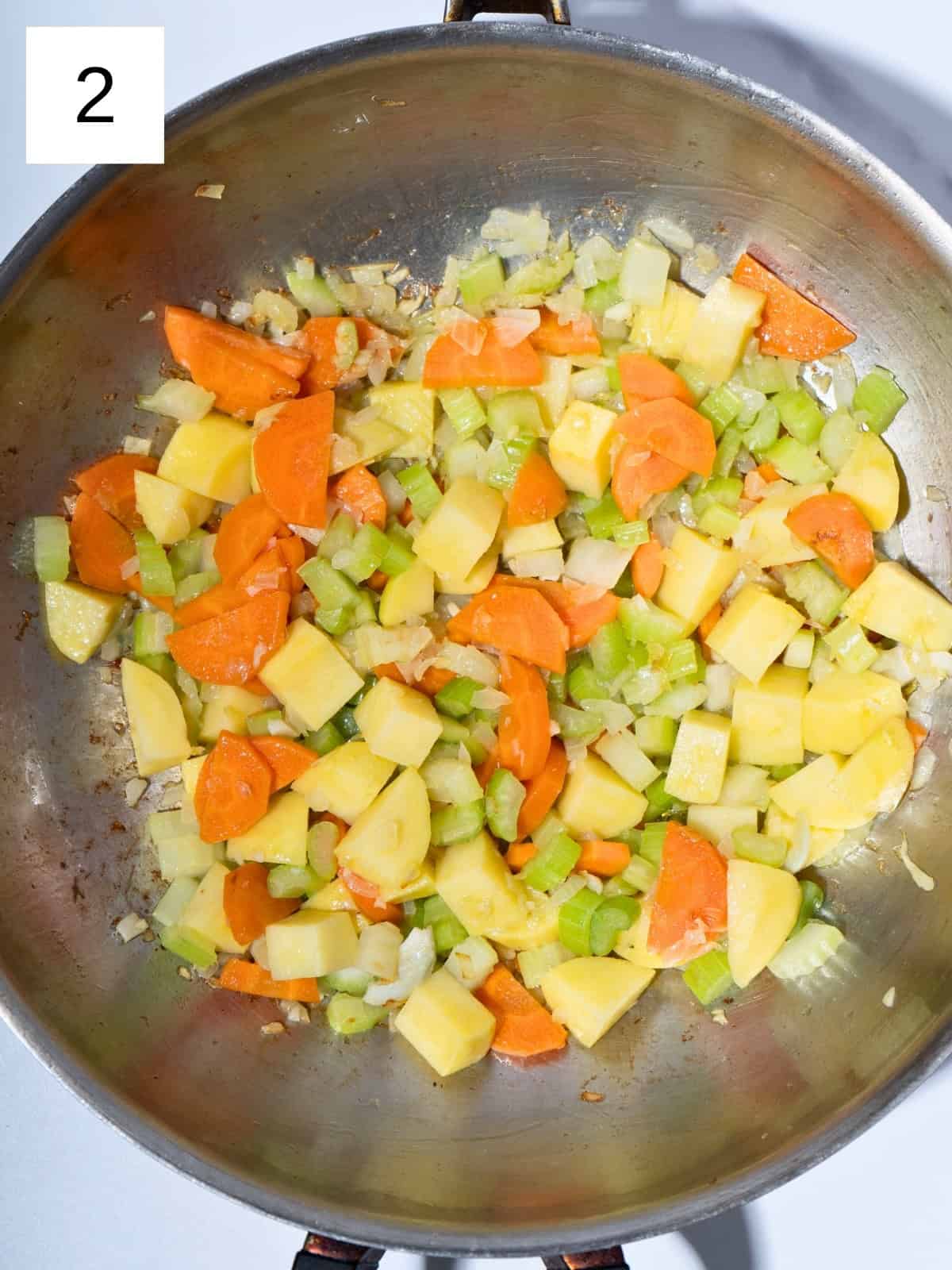 Chopped onion and garlic sautéing in a pot, with sliced carrots, diced potatoes, and chopped celery added to create the flavorful base of a hearty vegetable stew.