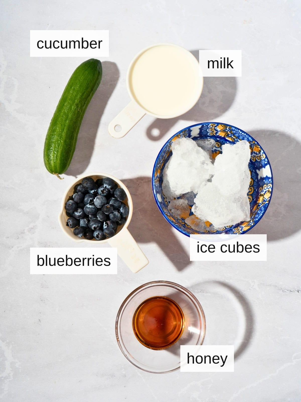 Ingredients for blueberry cucumber smoothie, including fresh cucumber, blueberries, milk, ice cubes, and honey.