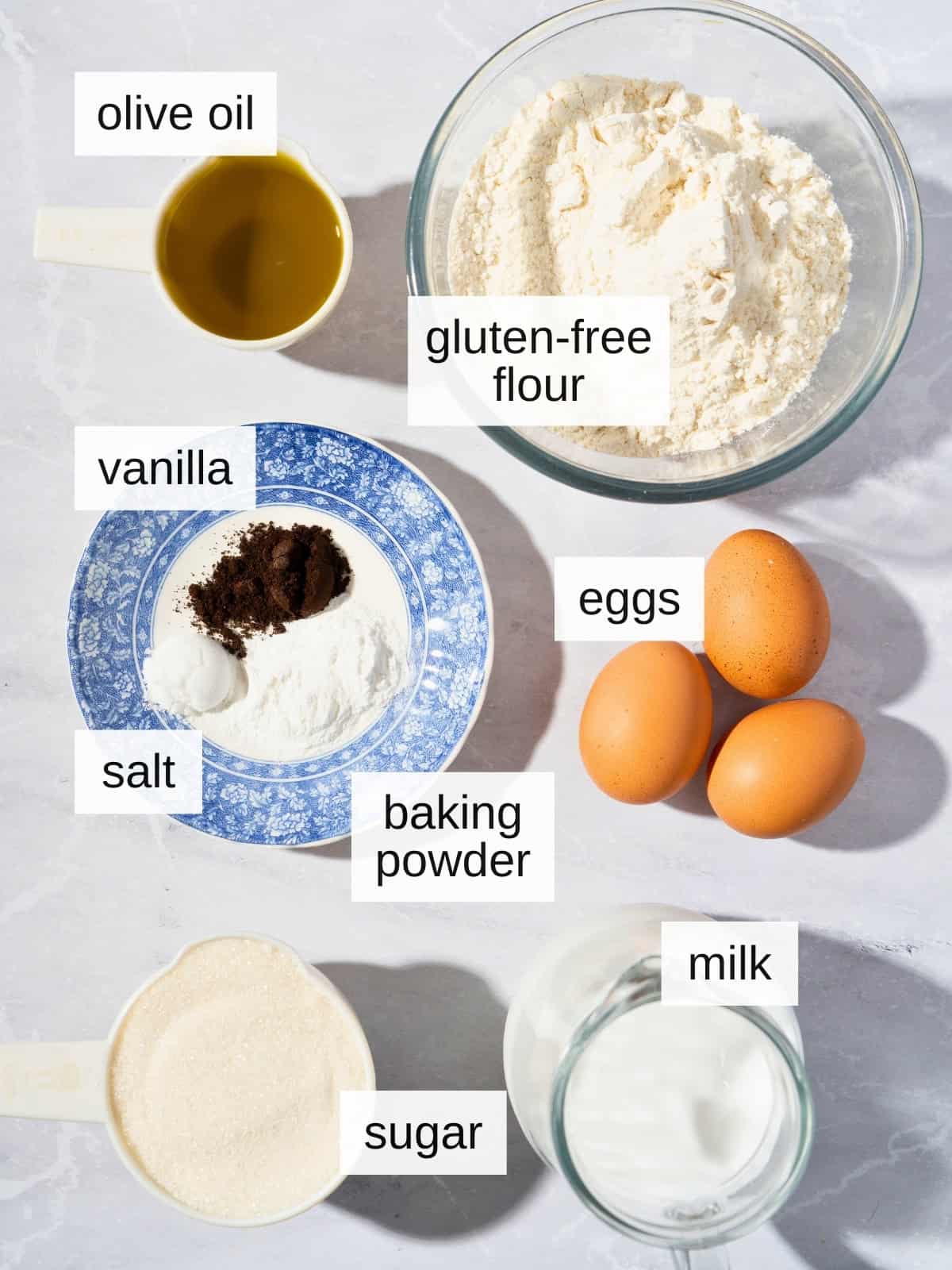 Ingredients for vanilla olive oil cake, including olive oil, gluten-free flour, vanilla, salt, baking powder, eggs, milk, and sugar.