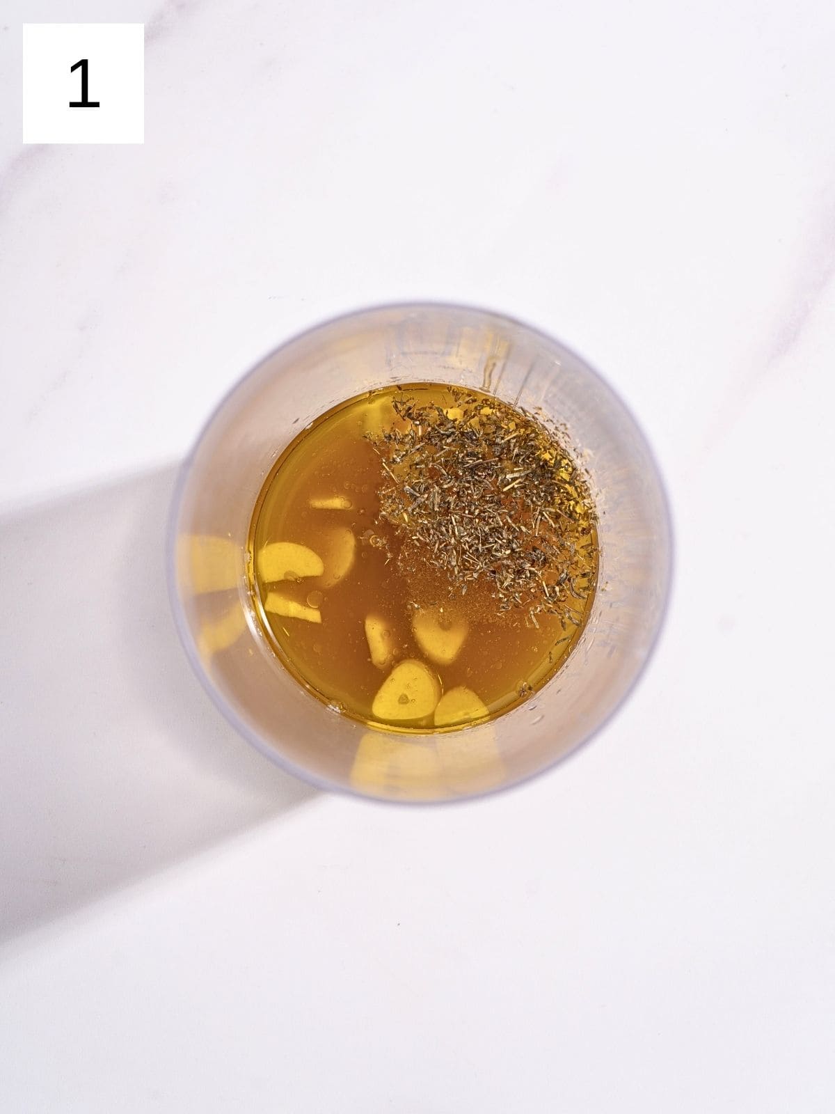A food processor bowl containing a mixture of olive oil, apple juice, minced garlic, fresh thyme leaves, and salt, ready to be blended into a flavorful dressing.