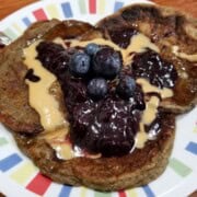 A stack of oat milk pancakes on a plate, topped with nut butter, fruit jam, and some fresh blueberries.