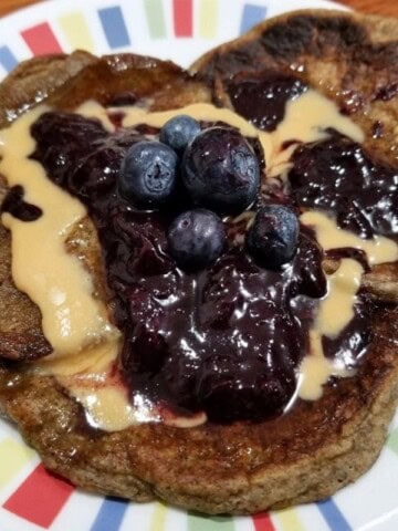A stack of oat milk pancakes on a plate, topped with nut butter, fruit jam, and some fresh blueberries.