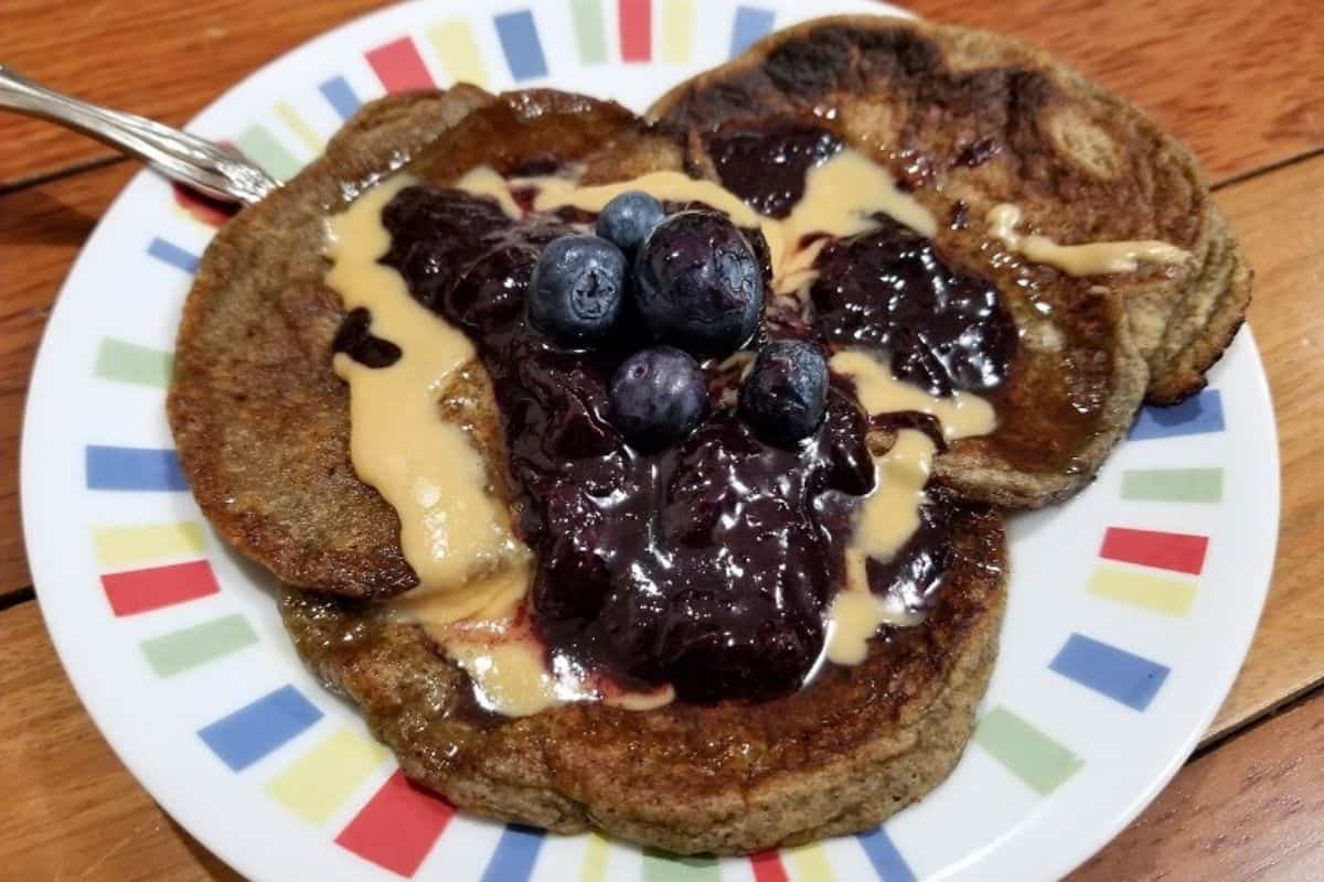 A stack of oat milk pancakes on a plate, topped with nut butter, fruit jam, and some fresh blueberries.