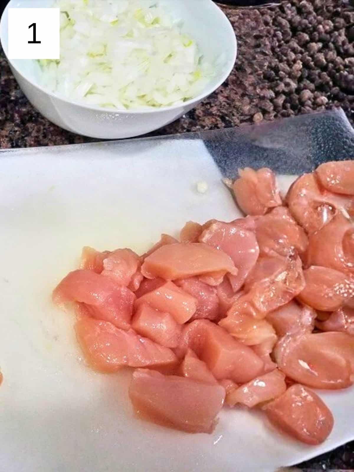 Chopped chicken breast on a chopping board, beside a bowl of chopped onions.